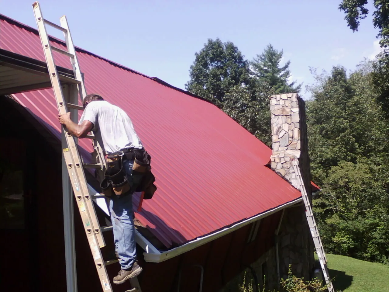 Roofer on a ladder beside a red metal roof and stone chimney — Soffit & Fascia Repair by Roofing Brothers in Ghent